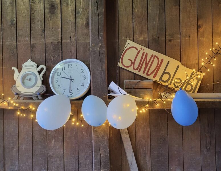 Eine Lichterkette, Uhr und Luftballons hängen an einer Holzwand, auf einem Pappschild steht Gündi West.
